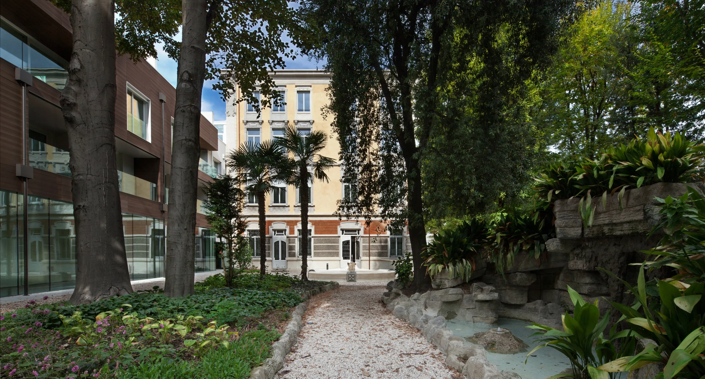 Sentiero ghiaioso tra alberi e piante in un giardino con fontana rocciosa, davanti a un edificio storico con facciata beige e dettagli bianchi.