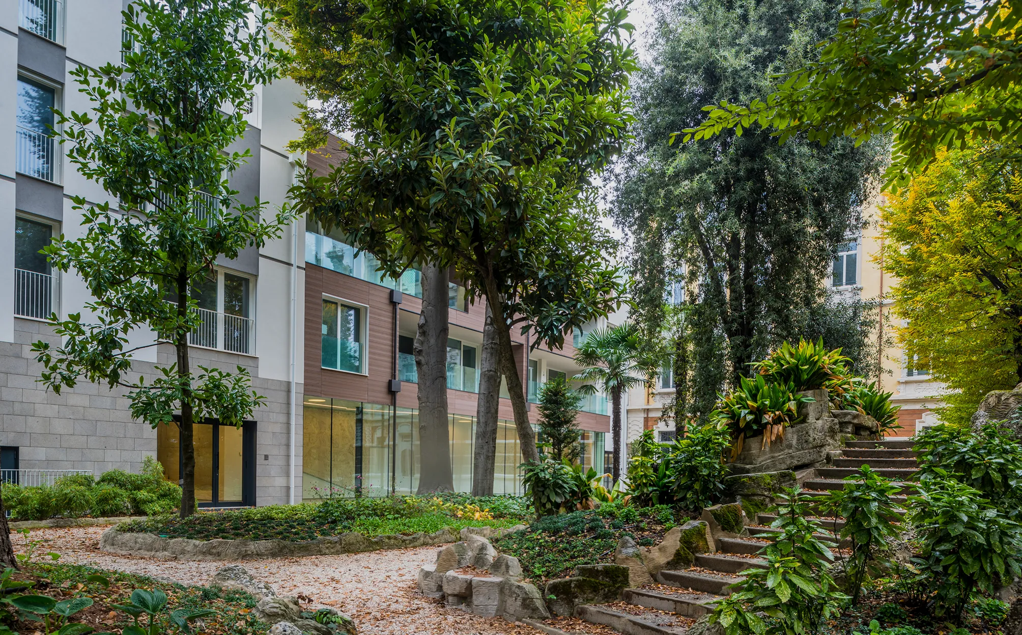 Giardino urbano rigoglioso con alberi, piante verdi e sentieri in ghiaia accanto a edifici moderni e antichi.