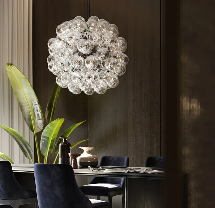 Lampada a sospensione composta da numerose lampadine di vetro sopra un tavolo scuro con sedie blu e piante verdi sullo sfondo.