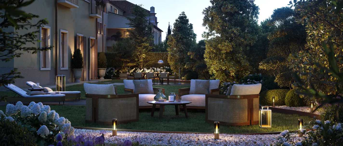 Giardino elegante di sera con sedie imbottite disposte attorno a un tavolino basso, illuminato da luci da terra e lampade soffuse, con vegetazione e piante curate intorno.