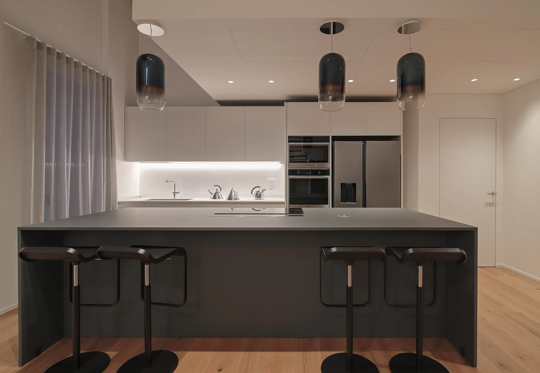 Cucina moderna minimalista con isola centrale grigia, quattro sgabelli neri, elettrodomestici integrati cromati e lampade sospese scure.