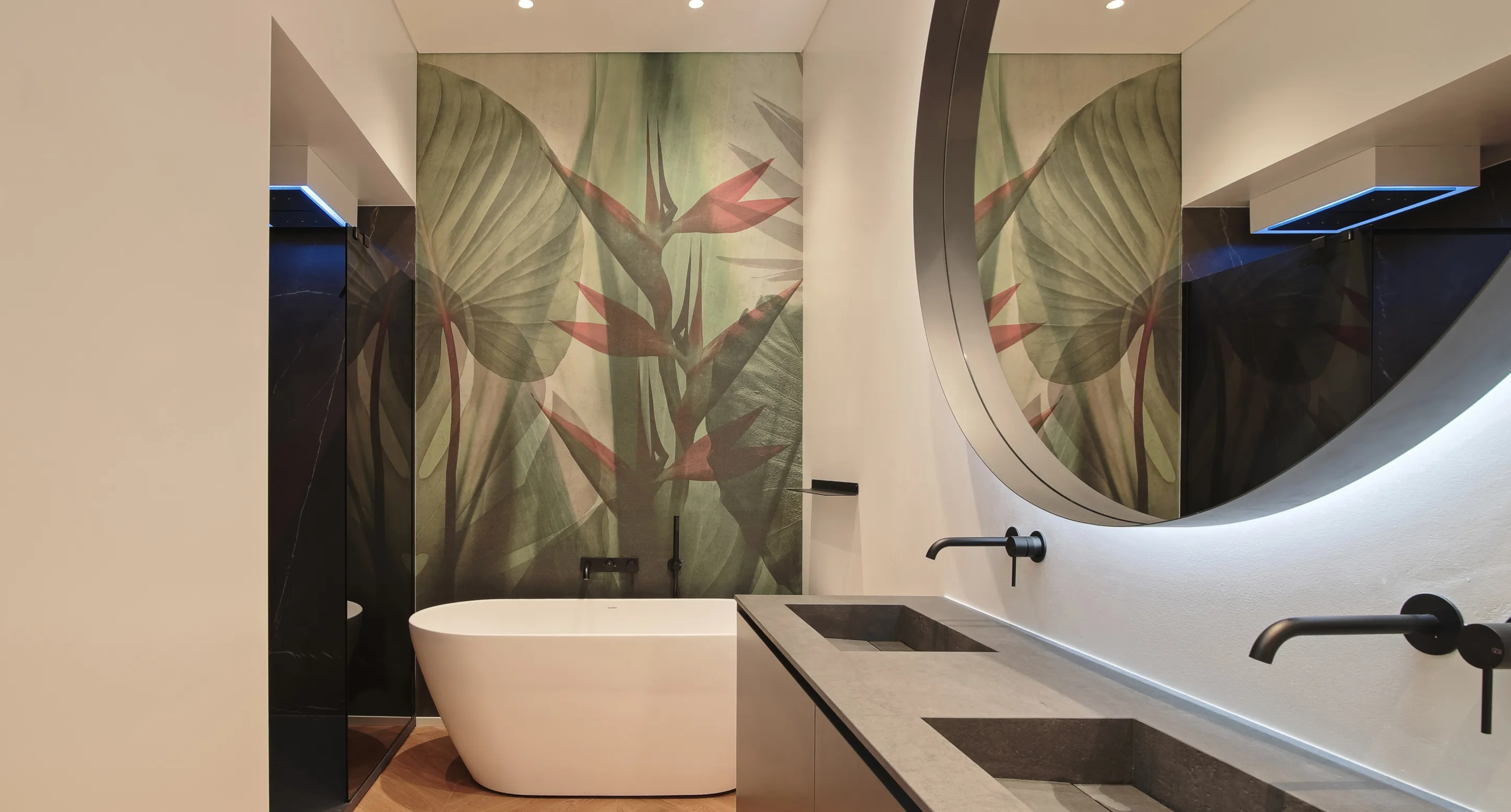 Bagno moderno con vasca bianca, doppio lavabo in pietra e grande specchio rotondo con parete decorata da murale botanico.