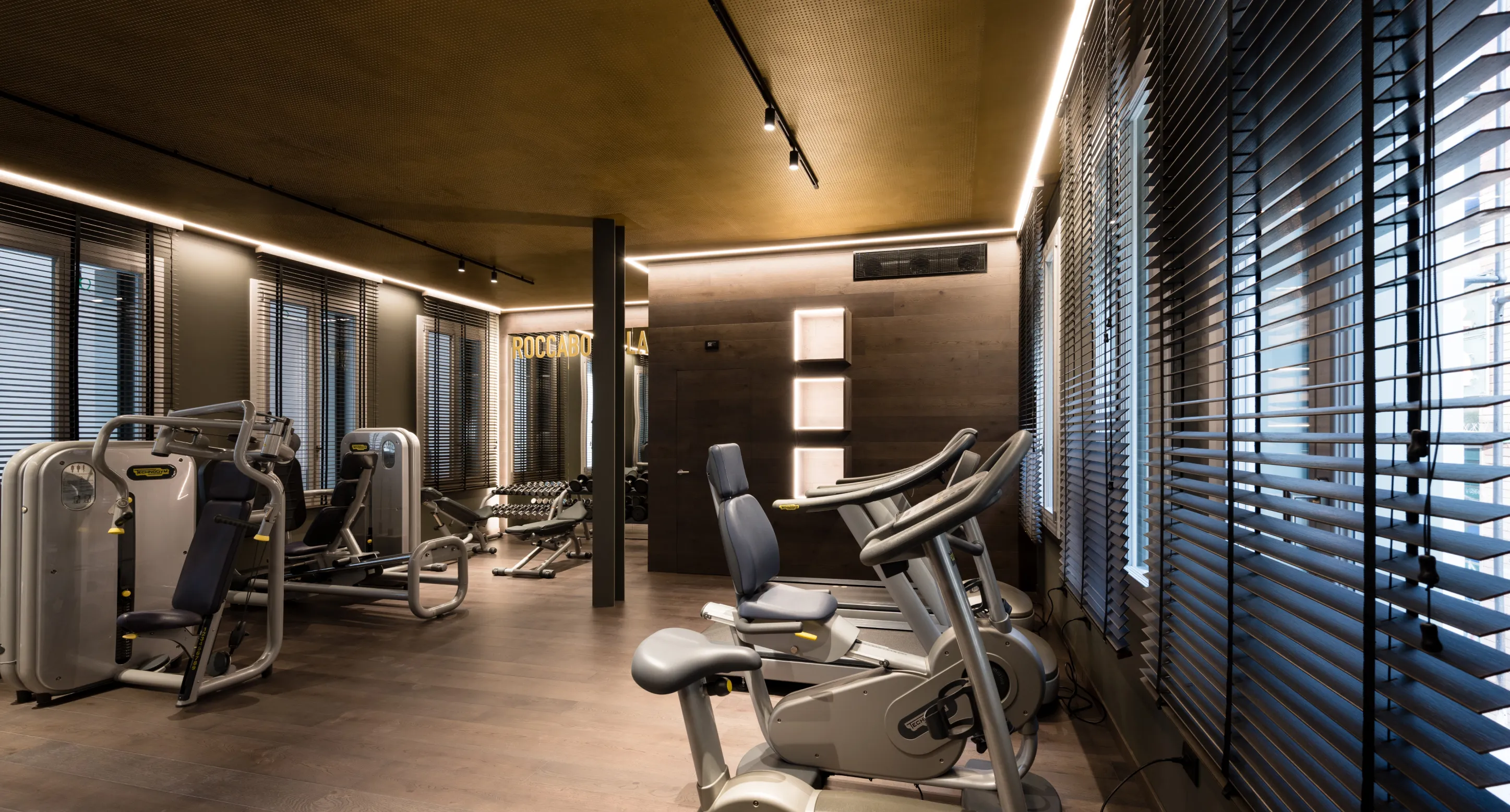 Palestra moderna con attrezzi fitness, cyclette e pesi liberi, illuminazione soffusa e grandi finestre con persiane.