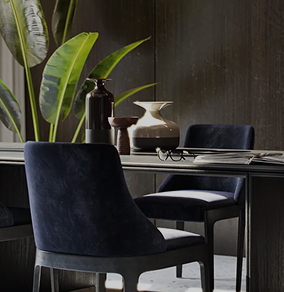 Modern dining table with dark upholstered chairs, decorative vases, and green leafy plant against a dark background.