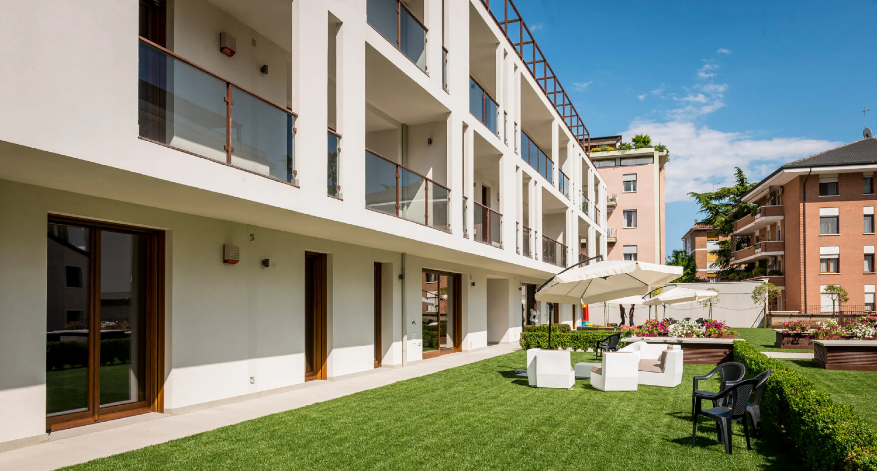Giardino ben curato con sedie bianche e nere sotto ombrelloni accanto a un moderno edificio residenziale con balconi in vetro.